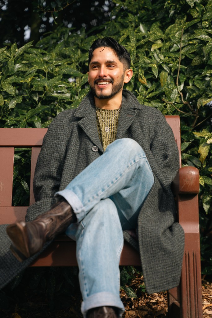 A man smiles while sitting on a bench on a sunny day. He wears a wool coat, yellow Mexican huipil, light washed jeans, and brown leather cowboy boots.